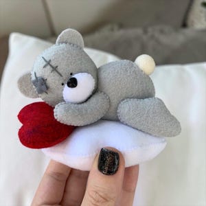 May include: A gray felt teddy bear toy with a red heart, resting on a white pillow. The bear has a black nose, white muzzle, and stitched details. The heart is a deep red color. The toy is handmade and has a cute, whimsical design.