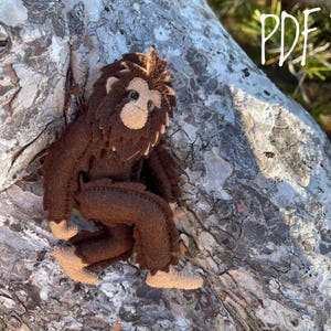 Puede incluir: Un juguete de peluche Bigfoot de fieltro marrón con una cara de color canela y ojos negros. El juguete está sentado en una rama de árbol.