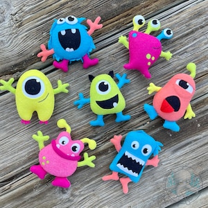 May include: A collection of colorful felt monster toys in various shapes and sizes. The monsters are in shades of blue, pink, yellow, and green, with googly eyes, mouths, and arms and legs. The toys are arranged on a wooden surface.