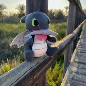 Peut inclure: Une peluche artisanale en forme de dragon, dans les tons de gris, blanc et rose, avec de grands yeux verts et de petites ailes. Le dragon est posé sur une rambarde en bois, avec un arrière-plan naturel de verdure.