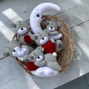 May include: A woven basket filled with handmade felt toys. The basket contains gray teddy bears, a white crescent moon, a red heart, and a white bunny. The bears have stitched details and patches. The toys are arranged on a white wooden surface.