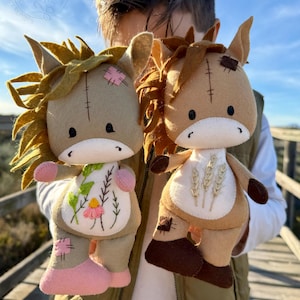 May include: Two handmade plush horse dolls. The dolls have tan bodies, brown manes, and stitched details. One doll has a floral design on its chest, while the other has wheat stalks. Both dolls have pink accents and patches.