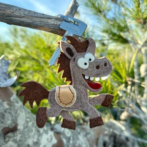 May include: A whimsical felt horse keychain in shades of brown and tan, with a saddle and a big smile. The keychain is attached to a set of keys, perfect for adding a touch of fun to your everyday carry.
