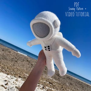 Pode incluir: Brinquedo de pelúcia de astronauta de feltro branco com detalhes pretos, sendo segurado no ar contra um fundo de céu azul e praia.  Padrão de costura PDF + TUTORIAL EM VÍDEO.