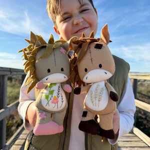 Puede incluir: Dos muñecos de peluche de caballo hechos a mano. Uno es de color beige con una flor rosa y pezuñas rosas, el otro es marrón con bordados de trigo y pezuñas marrones. Ambos muñecos tienen detalles cosidos y son sostenidos por una persona.