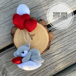 May include: A handmade gray teddy bear with a white face and a red heart-shaped pillow. The bear is holding two red heart-shaped balloons and a white balloon. The text "TEDDY BEAR WITH BALLOONS PDF PATTERN" is in a circle.