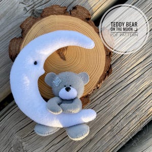 Puede incluir: Un oso de peluche gris abrazando una luna creciente blanca, sobre un fondo de rodaja de madera. El oso tiene un parche en la cabeza. La imagen incluye el texto "TEDDY BEAR ON THE MOON PDF PATTERN" en un diseño circular.