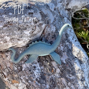 May include: PDF Sewing Pattern for a blue felt Nessie monster toy. The toy has a long neck, a small head with two black eyes, and a long tail. The toy is sitting on a piece of tree bark.