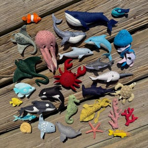 May include: A collection of handmade felt sea creatures, including a whale, dolphin, shark, octopus, crab, and various fish. The felt animals are in shades of blue, gray, red, and yellow, arranged on a wooden surface.
