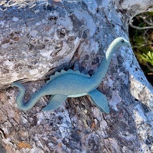 May include: A light blue felted dinosaur figurine with a long neck, a curved tail, and a row of small triangular spikes along its back. The creature has a smooth, soft texture and a single dark eye, positioned in a swimming pose with four fins.