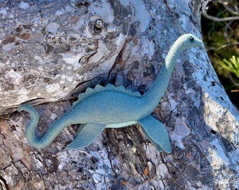 Könnte beinhalten: Eine hellblaue Dinosaurierfigur aus Filz mit langem Hals, geschwungenem Schwanz und einer Reihe kleiner dreieckiger Stacheln auf dem Rücken. Das Wesen hat eine glatte, weiche Textur, ein dunkles Auge und vier Flossen in einer Schwimmhaltung.