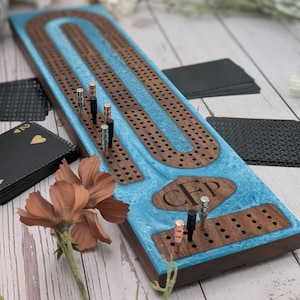 May include: A wooden cribbage board with a blue resin center and dark wood accents. The board features a track for pegging, with metal pegs in silver, black, and copper. A deck of cards and a flower are in the background.