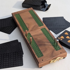 May include: A wooden cribbage board with green inlaid tracks and metal pegs, accompanied by several black playing cards with textured patterns and gold heart symbols, and a black card box with a gold-toned metal latch on the side.