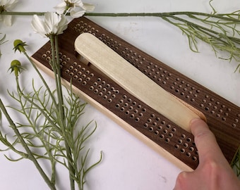 May include: A wooden cribbage board with a light brown wooden sliding piece. The board has a dark brown stain and is made of walnut wood. The board has a pattern of holes for pegs.