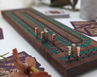 May include: A wooden cribbage board with a dark brown finish, teal and black inlaid tracks, and metal pegs in gold, silver, and copper colors. The board is rectangular with rounded ends. Playing cards are visible in the background.