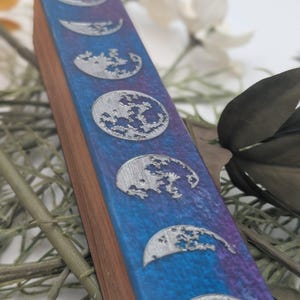 Peut inclure: Un porte-encens en bois avec une incrustation de résine bleue et violette. Le support présente des motifs de phases lunaires en métal argenté. Le bois a un grain naturel et la résine un effet marbré. Le design est sur le thème céleste.