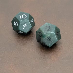 May include: Two dark green, multi-sided dice with white numbering and symbols. The dice are made of a speckled, semi-translucent material. One die shows the number 10, the other shows the number 2. The dice are resting on a brown surface.
