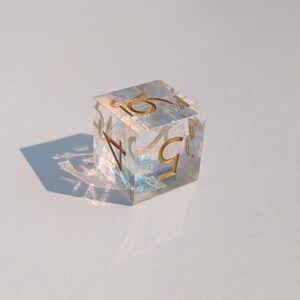 May include: A clear, iridescent resin die with gold-colored numbers. The die is a cube shape, with the numbers 1 through 6 visible on its faces. The die is casting a shadow on a white surface.