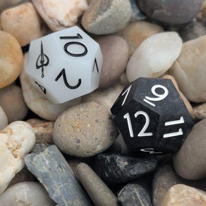 May include: Two twelve-sided dice, one white and one black, rest on a bed of smooth stones. The white die displays the number 10 and a symbol, while the black die shows the number 12. The dice are designed for tabletop gaming.