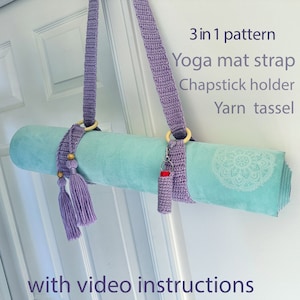 Peut inclure: Une sangle de tapis de yoga en crochet violet avec un anneau en bois, un porte-baume à lèvres assorti et un pompon en laine. La sangle est accrochée à une porte et un tapis de yoga est enroulé et sécurisé avec la sangle. Le texte "3 in 1 pattern" est écrit au-dessus de l'image. Le texte "Yoga mat strap, Chapstick holder, Yarn tassel" est écrit sous l'image. Le texte "with video instructions" est écrit sous le tapis de yoga.