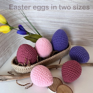 Può includere: Uova di Pasqua all'uncinetto in due dimensioni, rosa, viola e bordeaux, in un cesto intrecciato con carta triturata. Le uova sono disposte in un cesto con carta triturata. Il testo "Easter eggs in two sizes" è in alto nell'immagine.