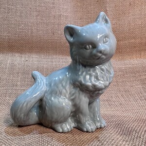 May include: A light blue ceramic cat figurine. The cat is sitting upright with its tail curled around its body. The figurine has detailed features, including ears, eyes, and fur texture. The cat is a solid color and has a glossy finish.