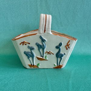 May include: A white ceramic planter with a handle shaped like a basket. The planter is decorated with blue and brown deer and a green and brown floral design.