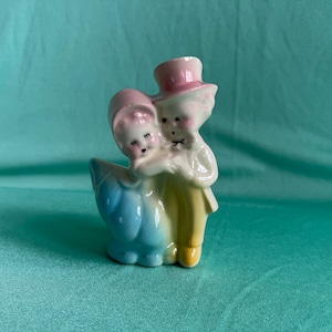 May include: A vintage ceramic planter shaped like a couple holding each other. The woman is wearing a blue and white dress and the man is wearing a yellow suit and a pink hat.
