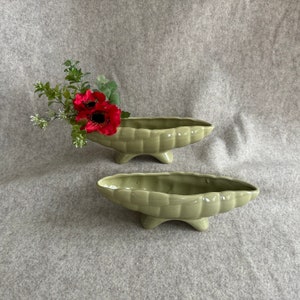 May include: Two green ceramic planters shaped like a pea pod with three legs each. The planters are on a gray textured surface. One planter has red silk flowers inside.
