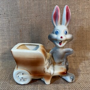 May include: A ceramic planter shaped like a rabbit pulling a wheelbarrow. The rabbit is gray with blue eyes and a red nose. The wheelbarrow is brown and white.