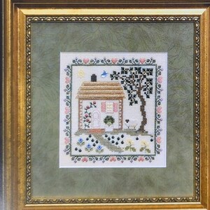 May include: A cross stitch pattern featuring a cottage with a tree and flowers. The pattern is framed in a gold frame and is accompanied by a quote: "Happy is the Home that shelters a Friend".