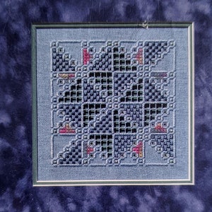 May include: Framed cross-stitch artwork featuring a geometric design in shades of blue, black, and red. The artwork is set within a dark blue mat and a green frame. The background includes daisies and greenery.