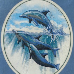 May include: A cross stitch kit featuring a design of three dolphins leaping out of the water. The kit includes 14 count white Aida fabric, embroidery thread, a needle, and instructions. The finished design measures 10 inches by 12 inches. The kit is packaged in a clear plastic bag with a black and white label that says "DIMENSIONS no count cross stitch" and "LEAPING FOR JOY designed by Peter Kull".
