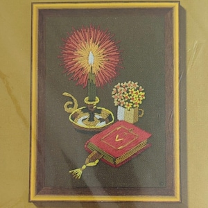 May include: A jiffy stitchery embroidery kit featuring a candlelight study design. The kit includes a printed design on homespun fabric, yarn, a needle, mounting board, and instructions. The design features a lit candle, a book, and flowers in a vase. The kit fits a standard 5 x 7 inch frame.