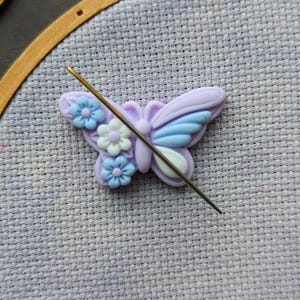May include: A purple butterfly with white flowers is attached to a needle in a wooden embroidery hoop. The hoop is on a purple fabric.