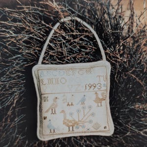 May include: A cross-stitch sampler with a handle, featuring peacock designs and the year 1993. The sampler is off-white with a cream-coloured handle, set against a backdrop of twigs and a wooden surface. The text "Peacock Spot Sampler" is handwritten at the top.