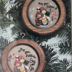 May include: Two cross-stitch patterns for holiday ornaments featuring a snowman and two children singing "Noel" with the text "May your Holiday Sing with Joy".