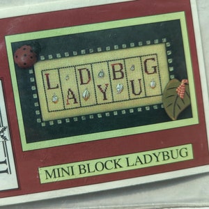 May include: A red and green cross stitch pattern card with a ladybug design. The card reads "Mini Block Ladybug" and features the words "L.D.B.G. A.Y.U."