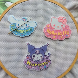 May include: Three embroidered patches featuring popular Sanrio characters: Cinnamoroll, Hello Kitty, and Kuromi. Each patch is on a white cloud with a colourful background and glitter accents. The patches are displayed on a wooden embroidery hoop.