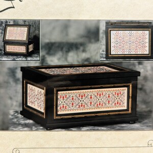 Puede incluir: Un cofre español negro con intrincados paneles estampados en rojo, blanco y dorado. El cofre tiene forma rectangular con tapa abatible y ribete dorado. El texto "Spanish Casket" es visible.
