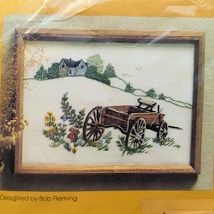 May include: A framed embroidery design featuring a brown wagon with a wooden wheel and a small house in the background. The design is titled "Old Wagon" and is designed by Bob Fleming.
