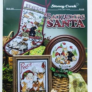 May include: A cross-stitch pattern booklet featuring a collection of backwoods Santa designs. The cover features a red stocking with a cross-stitch design of Santa and woodland animals, a round frame with a cross-stitch design of Santa and woodland animals, and a square frame with a cross-stitch design of Santa and woodland animals with the text "Peace on Earth".