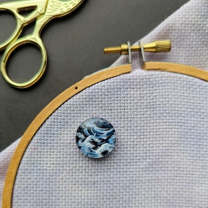 May include: A round, blue and white embroidery hoop with a close-up of a hand-painted design of a wave.