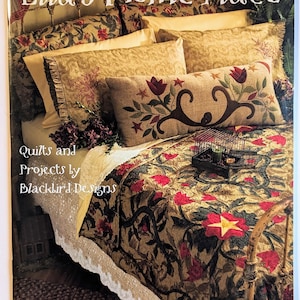Puede incluir: Una portada de libro titulada Ella's Home Place: Quilts and Projects by Blackbird Designs por Barb Adams & Alma Allen. Muestra una cama con una colcha floral, un cojín rectangular largo con bordados de flores y una pequeña jaula de alambre.