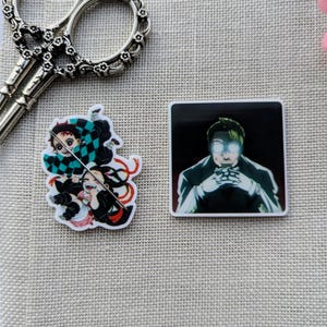 Könnte beinhalten: Zwei Anime-Pins und eine dekorative Schere liegen auf einem beigefarbenen Stoff. Ein Pin zeigt Charaktere mit schwarz-grünen Karomustern. Der andere Pin zeigt einen Charakter mit leuchtenden Augen. Die Schere ist silberfarben.