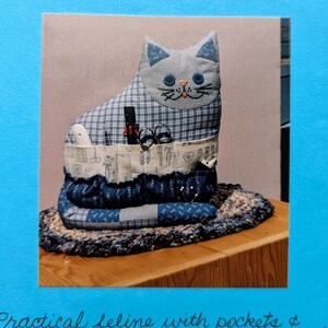 Peut inclure: Un organiseur de couture fait à la main en forme de chat, en tissu vichy bleu et blanc. Le chat a des traits de visage brodés et contient des fournitures de couture dans ses poches. L'organisateur est posé sur un tapis à motifs et une surface en bois.