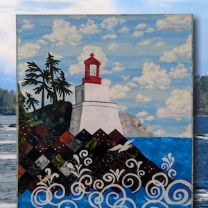 May include: A wall hanging titled WILD PACIFIC LIGHTHOUSE by Susan Teece, measuring 24 inches by 31 inches. The artwork depicts a white lighthouse with a red top, dark evergreen trees, and a rocky shore made of dark patterned fabric squares.