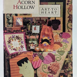 Può includere: Un'immagine colorata con vari oggetti trapuntati a tema autunnale. L'immagine include un cuscino con un disegno di fantasma, un asciugamano decorativo con un gatto nero e un oggetto a forma di Frankenstein. Il testo "Acorn Hollow" e "Art to Heart" sono visibili.