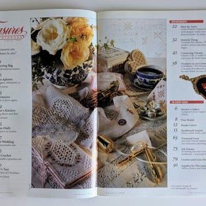 Op de afbeelding: Een tijdschriftpagina met een bloemstilleven, een kopje thee en borduurwerk. Het tijdschrift heet "Treasures in Needlework". De afbeelding bevat een lijst met artikelen en rubrieken.