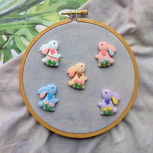 May include: Five colourful bunny shaped needle minders in a wooden embroidery hoop. The bunnies are pink, light blue, peach, purple, and white with embroidered details.